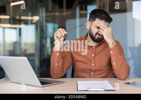 Ein junger Mann fühlt sich müde und hat schmerzhafte Kopfschmerzen, reibt sich unangenehm an den Schläfen, während er eine Brille hält, erlebt Burnout von der Arbeit und eine Belastung der Augen im Büro Stockfoto