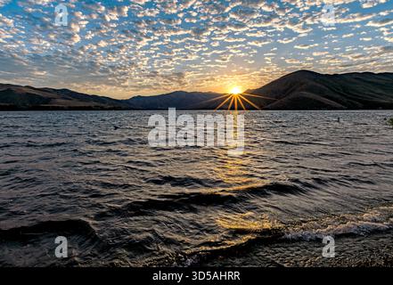 Sonnenstrahlen über dem Lucky Peak Reservoir, Idaho. Blauer Himmel mit aufgeschwollenen Wolken über der Sonne, die über dem Stausee aufgeht und Wellen am Ufer plätschern. Stockfoto
