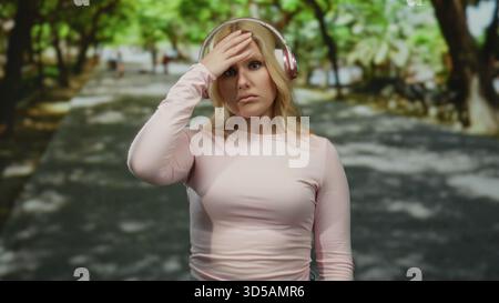 Frau, die im Park steht und Kopfhörer trägt, die Hand mit einem nachdenklichen Ausdruck hebt, umgeben von üppigen grünen Bäumen und natürlichem Licht, porträtiert Stockfoto