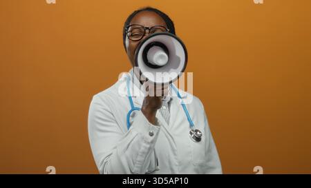 Frau Ärztin mit Stethoskop, die durch Megaphon vor orangem Hintergrund schreit, Begeisterung und Kommunikation ausdrückt. Stockfoto