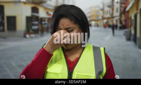Eine Frau, die tagsüber eine reflektierende Weste trägt und auf einer belebten Straße müde aussieht, deutet auf städtischen Stress und Müdigkeit hin. Stockfoto