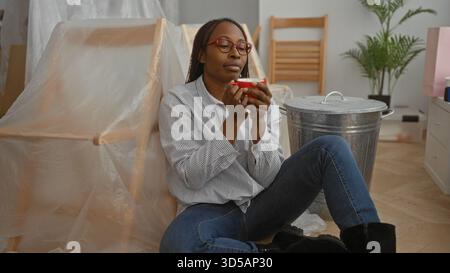 Frau, die sich in einem neuen Wohnraum entspannt und auf dem Boden sitzt, mit einer Tasse umgeben von unverpackten Möbeln, die mit Plastik bedeckt sind, und einer Pflanze im Hinterhaus Stockfoto