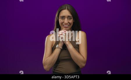 Junge hispanische Frau, die mit geschlossenen Augen und einem leichten Lächeln im lila Studio die Hände umschließt; hoffnungsvolle Dankbarkeit. Stockfoto
