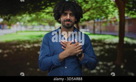 Mann mit lockigen Haaren, der die Hände über der Brust im Park nahe dem Gebäude hält, lächelt mit sanftem Ausdruck; aufrichtige Dankbarkeit. Stockfoto