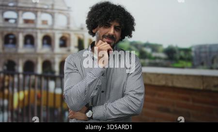 Mann mit lockigen Haaren und Hand am Kinn am kolosseum, lehnt sich auf ein Geländer und lächelt leicht; nachdenkliche Reflexion. Stockfoto