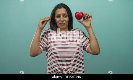 Junge hispanische Frau hält rotes Herz mit erhobener Hand im Studio vor türkisfarbenem Hintergrund; Skepsis. Stockfoto