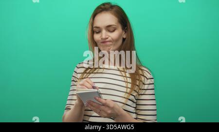Junge kaukasische Frau, die mit blauem Stift im grünen Atelier mit gestreiftem Hemd auf Notizblock schreibt; Besinnung. Stockfoto