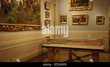 Innenraum der Museumsgalerie mit gerahmten Gemälden und einem verglasten Vitrinentisch in sanftem, unscharfem Bokeh; Hintergrund im Hintergrund mit ruhiger Atmosphäre im Copyspace. Stockfoto