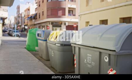 Eine Reihe von grauen und grünen Recyclingbehältern und geparkten Autos, die in flachem Bokeh auf einer sonnendurchfluteten Stadtstraße mit weichen, unscharfen Gebäuden und Bürgersteig gefangen wurden; bac Stockfoto