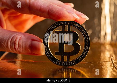 Nahaufnahme der Hand, die physische Gold-Bitcoin-Münze auf reflektierender goldener Oberfläche hält Stockfoto