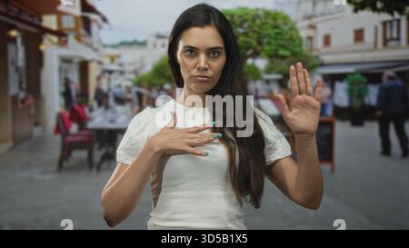 Frau junge hispanische Hand auf der Brust und erhöhte Handstopp Geste im Straßencafé mit weißem T-Shirt; Eid feierlich. Stockfoto
