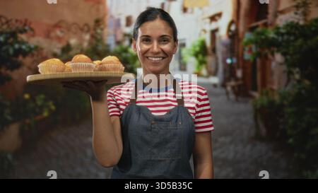 Frau mit hölzernem Tablett von Muffins auf erhobener Hand in einem kopfsteingepflasterten Straßenmarkt; Gastfreundschaft kleines Geschäft. Stockfoto