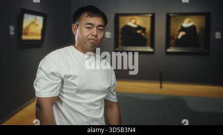 Junger chinesischer Mann in weißem T-Shirt steht still und starrt auf gerahmte Gemälde an der Wand im schwach beleuchteten Galeriebau; Nachsicht. Stockfoto