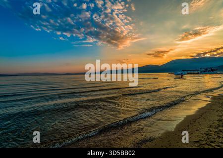 Goldener Sonnenaufgang über dem See Ohrid mit sanften Wellen und entferntem Pier in Pogradec, Albanien Stockfoto