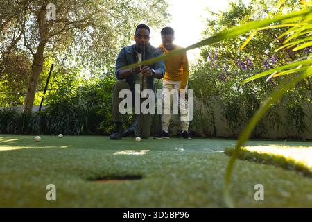 Afroamerikanischer Vater und Sohn spielen Minigolf auf dem Rasen auf dem Golfplatz im Freien mit verstreuten Bällen Stockfoto
