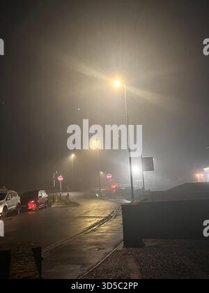Nächtliche urbane Szene mit dichtem Nebel, hellen Straßenlaternen und reflektierendem nassem Asphalt auf einer ruhigen Straße in Hull, England. Stockfoto