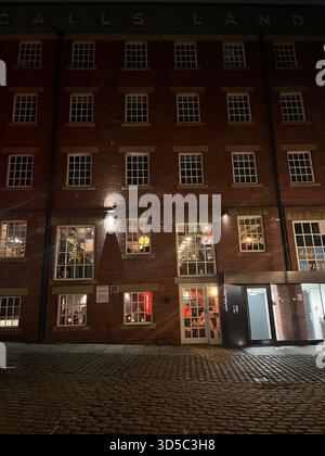 Historisches rotes Backsteingebäude mit beleuchteten Fenstern bei Nacht in Leeds, England. Atmosphärische urbane Szene mit Kopfsteinpflasterstraße und warmer Inneneinrichtung Stockfoto