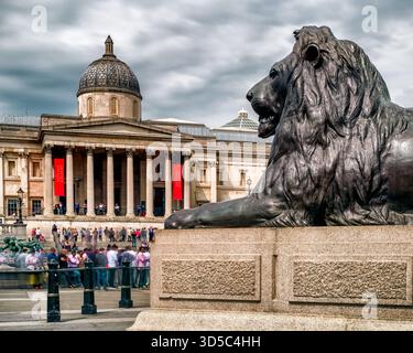London, Großbritannien - 14. Mai 2018: Löwenstatue am Trafalgar Square mit der National Gallery im Hintergrund an einem bewölkten Tag im Zentrum Londons. Stockfoto