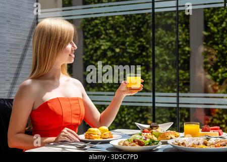 Frau in orangefarbenem Kleid, die Frühstück mit Saft auf sonnigem Freien genießt Stockfoto
