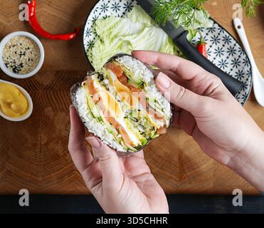 Ein kreativer Sushi-Burger mit Lachs, Spiegelei, Käse, Gurke und Avocado in den Händen einer Frau. Stockfoto
