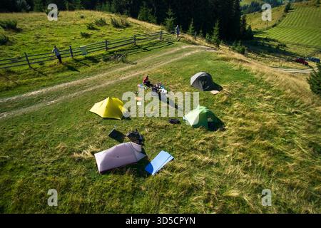 Campingplatz aus der Vogelperspektive in den Bergen, mit mehreren bunten Zelten auf einer grasbewachsenen Rodung. Camper entspannen unter klarem blauen Himmel, umgeben von hohen immergrünen und Panoramablick auf entfernte Gipfel. Stockfoto