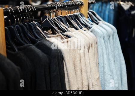 Eine Reihe gemütlicher Strickpullover in Schwarz, Beige und Hellblau, die an schwarzen Kunststoffbügeln an einer Schiene in einem Bekleidungsgeschäft hängen. Hochwertige Fotos Stockfoto