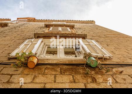 Blick nach oben auf ein mediterranes Steinhaus mit offenen weißen Fensterläden und kleinen Topfpflanzen unter dem Fenster. Die rustikale Architektur und das warme Licht spiegeln den Charme der südlichen Küstenstädte wider. Stockfoto