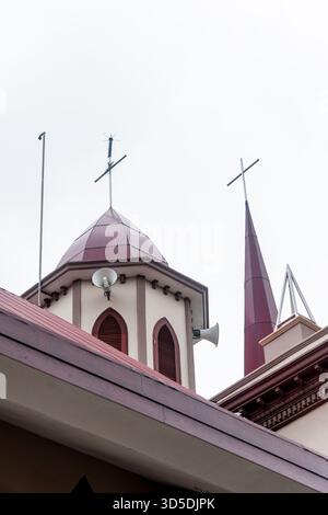 Eine architektonische Detailaufnahme eines Kirchengebäudes mit kastanienbraunen Dächern, zwei Türmen mit Kreuzen und Lautsprechern vor einem weißen bedeckten Himmel. Stockfoto