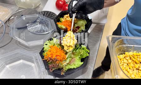 Der Koch bereitet frischen Gemüsesalat mit geriebenen Karotten, Rotkohl und Salat in schwarzen Schüsseln zu und trägt schwarze Handschuhe in der professionellen Küche Stockfoto
