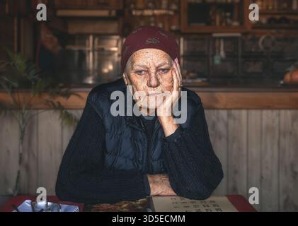 Eine 86-jährige Frau sitzt an einem Tisch in einem Landhaus und sieht nachdenklich aus Stockfoto