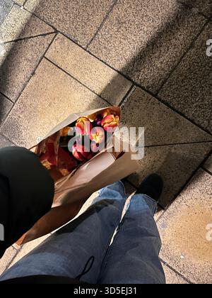 Eine Person, die auf einer gepflasterten Straße spaziert und Einkaufstaschen mit Tulpen hält. Die Szene zeigt einen alltäglichen urbanen Moment in Hull, England Stockfoto
