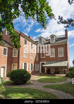 Die Seite des historischen Read House, ein großes rotes Backsteingebäude aus der Kolonialzeit mit weißen Fenstern, eingerahmt von üppig grünem Laub und blauem Himmel in New Castle Stockfoto