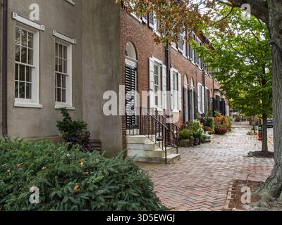 Eine bezaubernde Reihe von historischen roten Backsteinhäusern mit weißen Fensterläden entlang einer kopfsteingepflasterten Straße und einem Bürgersteig mit Herbstlaub in New Castle, Delaw Stockfoto