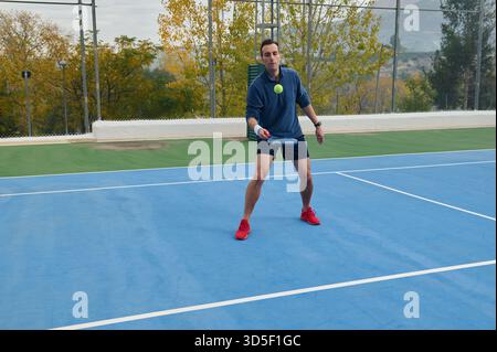 Ein männlicher Spieler in blauem Oberteil und roten Schuhen bereitet sich darauf vor, auf einem hellen Outdoor-Platz mit Bäumen und einem Zaun im Hintergrund einen Paddeltennisball zu schlagen. Stockfoto