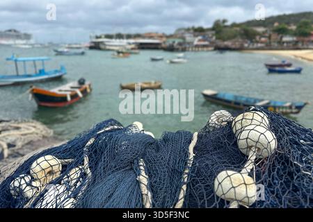 Armação dos Búzios, Rio de Janeiro, Brasilien. Fischernetz mit weißen Schwimmern im Vordergrund, mit Blick auf einen kleinen Hafen an einem bewölkten Tag. Stockfoto