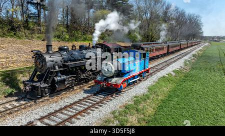 Paradise, Pennsylvania, 17. April 2025 - zwei Dampfzüge fahren an klaren Tagen parallel, umgeben von grünen Feldern und Bäumen. Ein Zug ist klassisch schwarz, der andere in leuchtendem Blau. Stockfoto