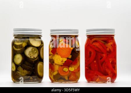 Drei Einmachgläser mit verschiedenen eingelegten Gemüsesorten stehen vor einem weißen Hintergrund. Die Gläser enthalten Gurkenscheiben, Karotten, Blumenkohl und Paprika und zeigen lebendige Farben und Texturen. Stockfoto
