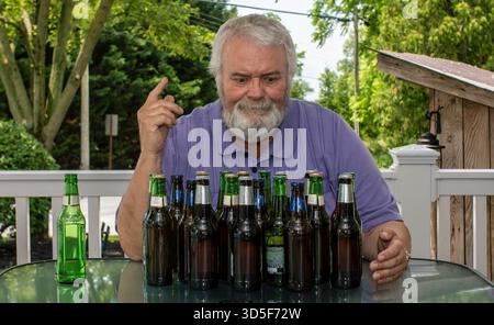 Ein Mann mit grauem Bart sitzt an einem Tisch im Garten und zählt leere Bierflaschen. Die Sonne scheint hell, während er einen entspannenden Nachmittag inmitten von Grün genießt. Stockfoto