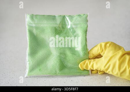 Gelbe Hand mit Handschuhen, die grünes Reinigungstuch in Plastiktüte auf einer hellen Oberfläche hält Stockfoto