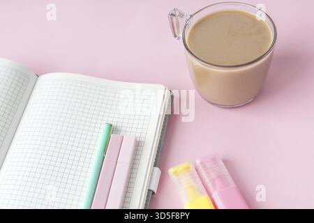 Ein offenes Notizbuch mit Pastellstiften, Textmarkern und einer Tasse Kaffee auf rosafarbenem Hintergrund. Die Arbeitsbereichseinstellung spiegelt Fokus und Kreativität wider Stockfoto