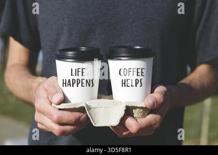 Eine Person in einem dunklen Hemd, die eine Kaffeetasse mit zwei Tassen zum Mitnehmen hält, die motivierende Zitate enthalten: Das Leben geschieht und Kaffee hilft. Begriff CAF Stockfoto