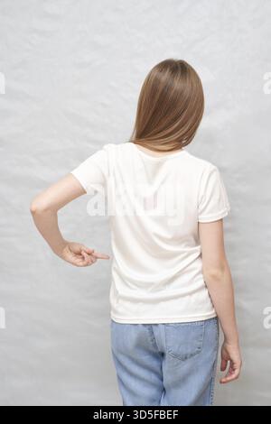 Junge weiße Frau in weißem T-Shirt und Jeans, die vor hellgrauem Hintergrund nach hinten zeigen Stockfoto