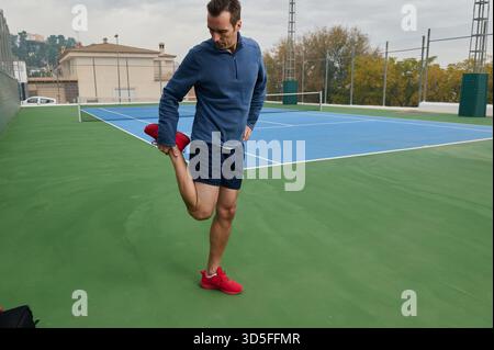 Ein Mann in blauem Oberteil und marineblauen Shorts streckt sein Bein auf einem blauen Tennisplatz und bereitet sich auf das Training vor. Outdoor-Umgebung, Fitness und Fokus werden betont Stockfoto
