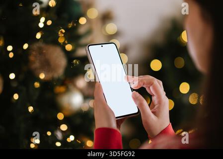 Nahaufnahme einer Frau mit Smartphone mit leerem weißen Bildschirm in der Nähe des Weihnachtsbaums mit goldenen Festlichtern Stockfoto