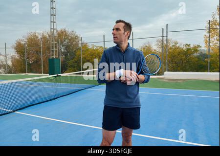 Ein Mann in blauem Pullover und Shorts steht auf einem blauen Tennisplatz, hält Schläger und Ball und blickt weg. Außenanlage mit Zaun, Bäumen und cl Stockfoto