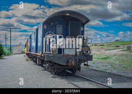 Ein alter blauer Zugwagen befindet sich auf Gleisen in ländlicher Umgebung, mit einem kleinen Gebäude und grünen Feldern in der Nähe Stockfoto