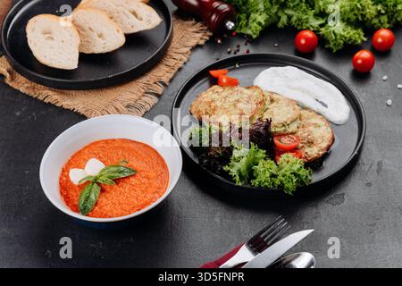 Tomatensuppe und Zucchini-Krapfen mit Joghurtsauce, Salat und Brot auf dunklem Tisch Stockfoto
