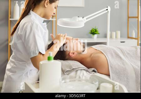Mann, der friedlich auf dem Spa-Bett liegt, professionelle Gesichtsbehandlung, weibliche Kosmetikerin sanft massiert Stockfoto