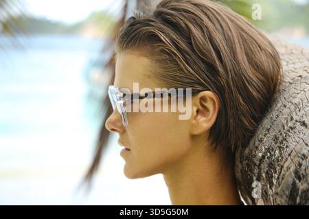 Kopfaufnahme einer schönen Frau mit metallischer Goldfarbe auf ihrem Gesicht und lehnt sich an die Palme. High-Fashion-Ästhetik im Urlaub Stockfoto