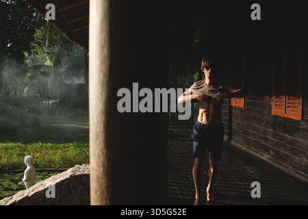 Mann, der auf einer dunklen Holzterrasse steht, beleuchtet von einem raffinierten Licht, das seine sportliche Note unterstreicht, während er ein T-Shirt anzieht. Stockfoto
